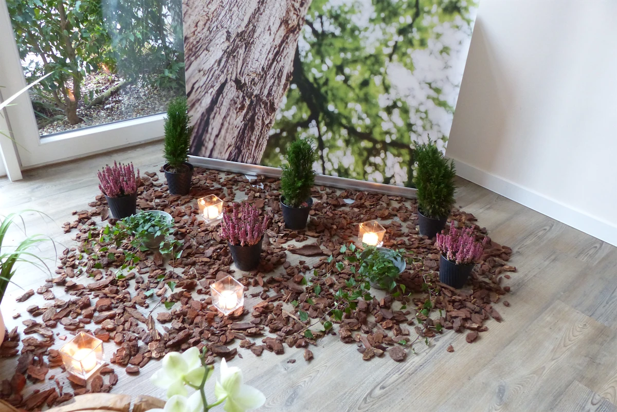 Fünfköpfige Familie mit drei Kindern, zwei Erwachsene, bei einem jungen Baum im Garten. Blumen, Pflanzen und Haus im Hintergrund sichtbar.